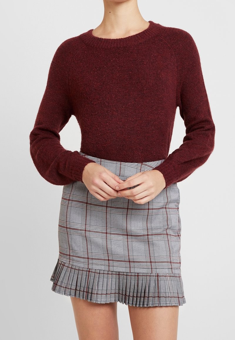 Burgunderrød strikket sweater med lange ærmer parret med en grå ternet mini-sokke med plisset kant og røde detaljer.