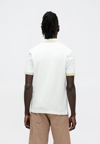 Fehér rövid ujjú póló világoszöld gallérral és ujjdíszítéssel. Az anyag sima és texturált hatású.