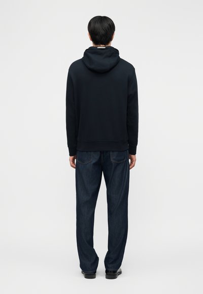 Άτομο που φοράει μαύρο φούτερ με κουκούλα, σκούρα μπλε τζιν και μαύρα παπούτσια, στέκεται με την πλάτη προς τα εμπρός σε απλό ανοιχτό φόντο.