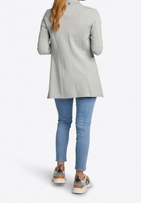 Vrouw met lang haar, gekleed in een lange grijze blazer, blauwe skinny jeans en witte sneakers met oranje en bruine accenten, staand met haar rug naar ons toegekeerd.