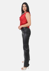 Vrouw met lang, donker haar, in een mouwloze rode top, zwarte leren broek en witte hoge hakken, staande in profiel.