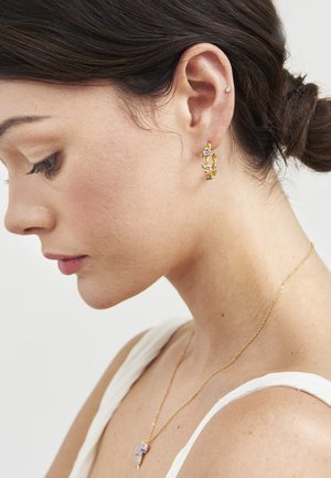 Pruunides kuklases juustega naine, kes kannab kuldseid lillemotiiviga rõngaskõrvarõngaid ja väikese ripatsiga kuldset kaelakeed, nähtud profiililt tavalise tausta ees.