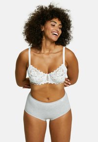 Donna sorridente con capelli ricci che indossa un reggiseno di pizzo bianco e mutande di cotone grigio chiaro a vita alta, in piedi davanti a uno sfondo bianco.