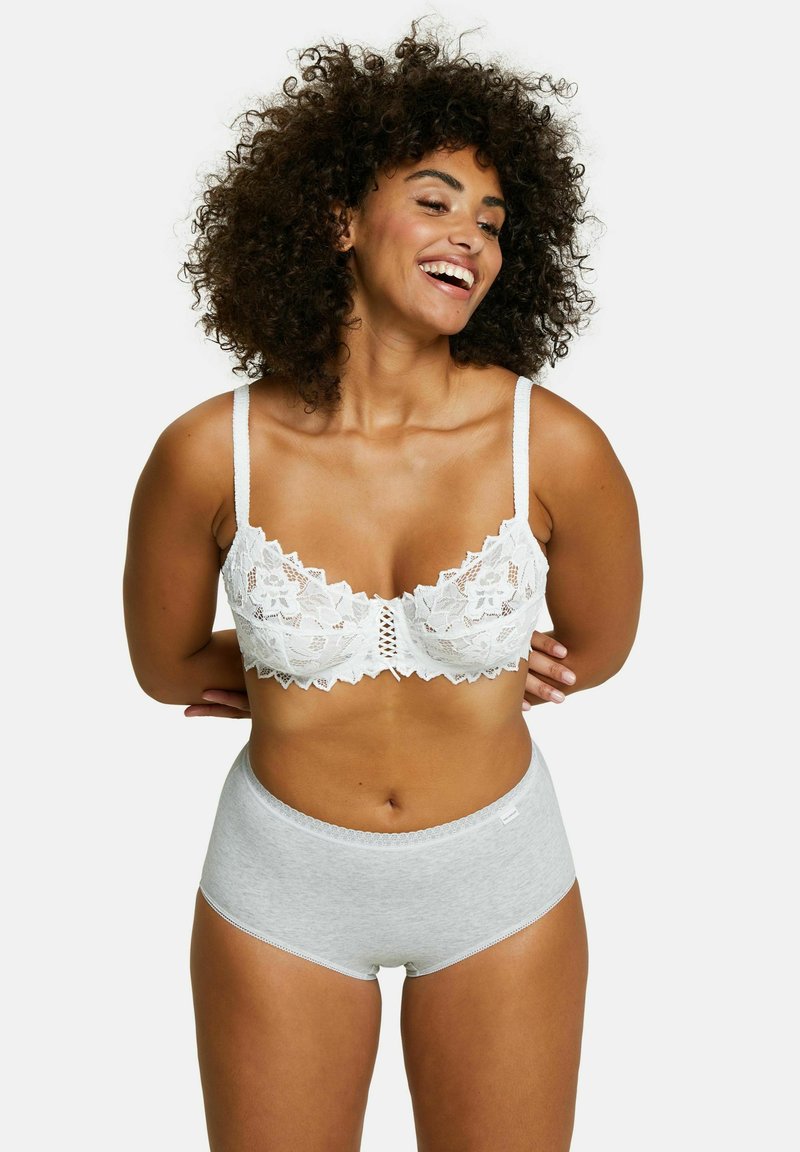 Donna sorridente con capelli ricci che indossa un reggiseno di pizzo bianco e mutande di cotone grigio chiaro a vita alta, in piedi davanti a uno sfondo bianco.