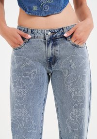 Jeans in denim blu chiaro con design di teschi argentati lungo le gambe, stile a cinque tasche, chiusura con bottone e leggera texture sbiadita.