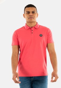 Polo shirt corail avec un patch logo noir, patte de boutonnage à trois boutons, et un accent blanc sur le col. Manches courtes, texture lisse. Coupe décontractée.