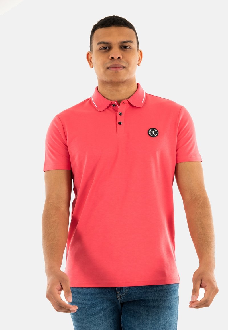 Polo shirt corail avec un patch logo noir, patte de boutonnage à trois boutons, et un accent blanc sur le col. Manches courtes, texture lisse. Coupe décontractée.