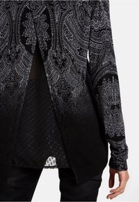 Schwarz-weißer Cardigan mit komplexem Paisley-Muster. Das Material hat eine strukturierte Oberfläche, die einheitliche und gemusterte Abschnitte verbindet.