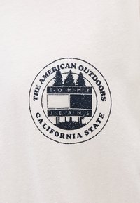 Bela tkanina vsebuje okrogel modri logotip z napisom "THE AMERICAN OUTDOORS" in "CALIFORNIA STATE", ob stiliziranih drevesih ter napisom "TOMMY JEANS".