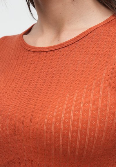 Τοπ σε χρώμα σκουριάς με ριμπ ύφανση και στρογγυλή λαιμόκοψη, που διαθέτει διακριτικά υφασμένα μοτίβα και απαλό ύφασμα. Κοντινή λήψη που αναδεικνύει τις λεπτομέρειες στις ραφές.