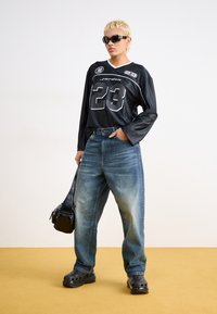 Svart langermet topp med "23" grafikk, blå baggy denimjeans, svarte tresko og en liten svart skulderveske. Flat tekstur og avslappet passform.