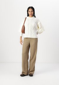 Maglione bianco a treccia con maniche ampie, abbinato a pantaloni beige a gamba larga e una borsa marrone. Scarpe nere semplici completano l'outfit.