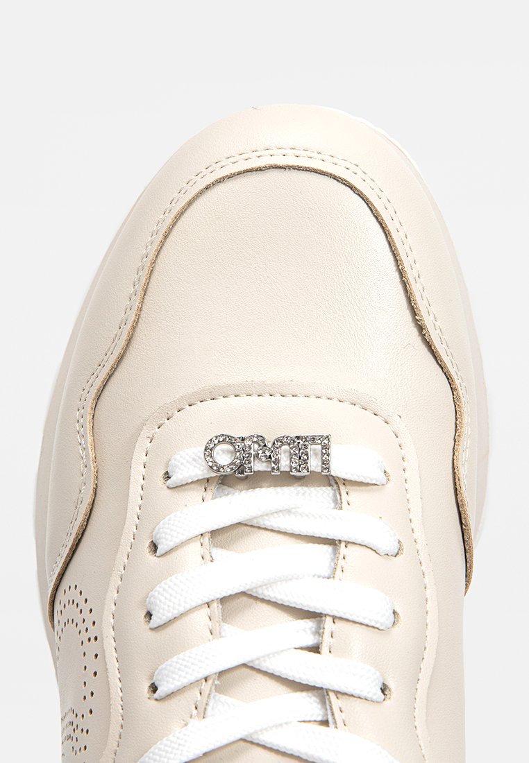 Zapatilla de cuero beige con cordones blancos, detalles perforados y acento de logo metálico. Punta redondeada y costuras en contraste.
