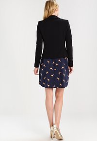 Musta istuva bleiseri, jossa on räätälöity siluetti, päällä laivastonsininen mekko, jossa on flamingokuvio. Hame on hieman liehuva; metallinhohtoiset korkokengät viimeistelevät asun.