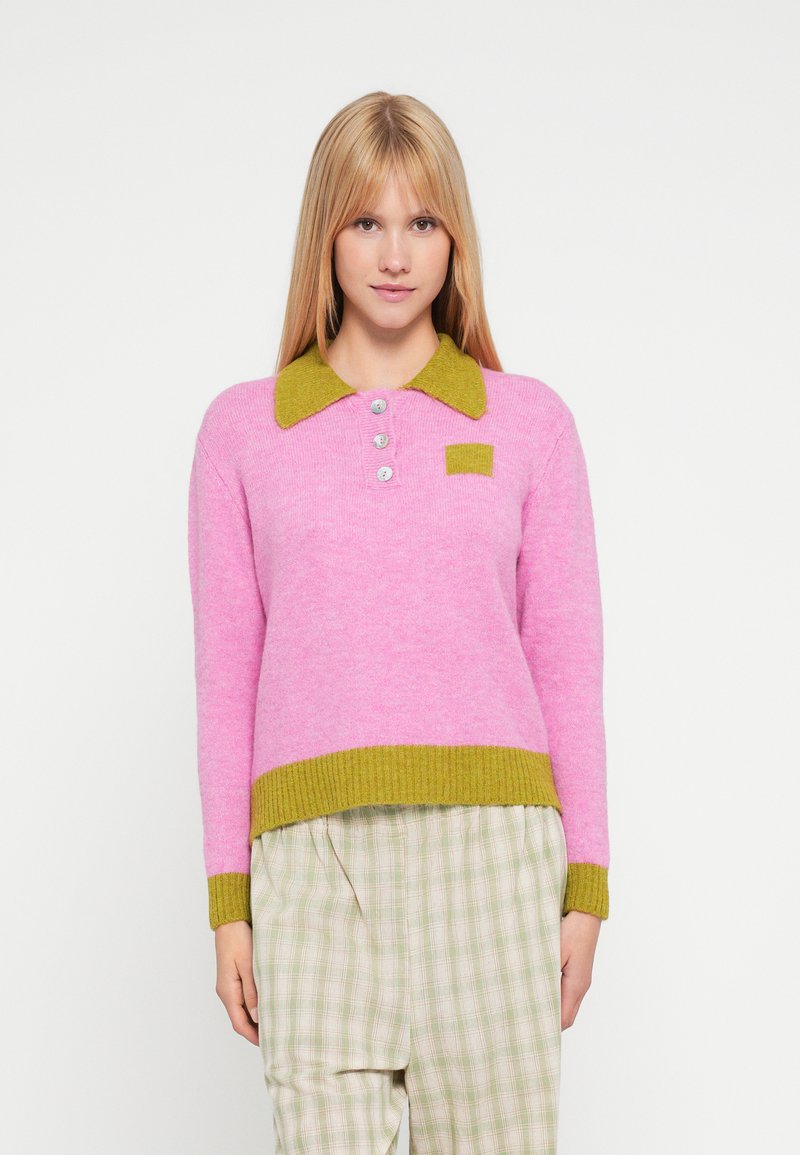 Maglione rosa lavorato a maglia con collo verde, tre bottoni e tasca applicata. Abbinato a pantaloni a quadri chiari e larghi.