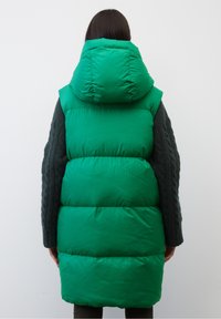 Grøn puffervest med høj krave og stor hætte, der har quiltede sektioner og en blank overflade, som er parret med teksturerede mørke ærmer.