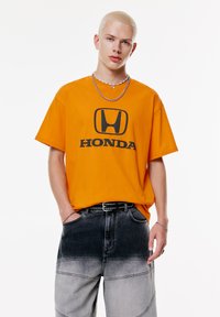 Orange bomulds t-shirt med sort "HONDA" logo på brystet, parret med grå ombre denim bukser. Modellen bærer lagdelte sølvkæder og et sort bælte.
