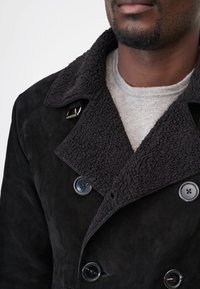 Sort ruskindsfrakke med en tekstureret, pelset krave, dobbeltradet front og to knapper. Har en justerbar metalspænde detalje.