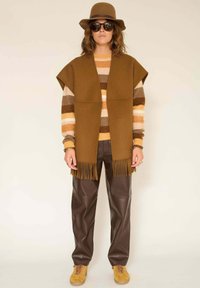 Gilet à franges marron sur un pull rayé dans des tons d'orange et de marron, associé à un pantalon en cuir marron foncé et des baskets jaunes.