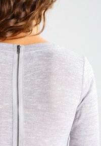 Top grigio a maglia con dettaglio della cerniera sul retro e un motivo testurizzato, finitura liscia e scollatura arrotondata.