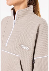 Beige halv-draget genser laget av teksturert stoff, med en hvit glidelås og kant. Viser en fremtredende "PEGADOR"-label.