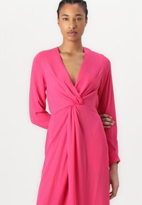 Pink long-sleeve dress with a deep V-neck and gathered waist detailing, made of soft, smooth fabric, featuring draped elements for flow.