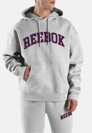 Mann trägt hellgrauen Reebok-Hoodie und passende Jogginghose mit großem marineblau-rotem Reebok-Logo auf Brust und Oberschenkel.