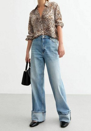 Person iført skjorte med leopardprint, lyseblå jeans med vide ben og opbuk, sorte spidse sko, der holder en sort håndtaske ved siden.