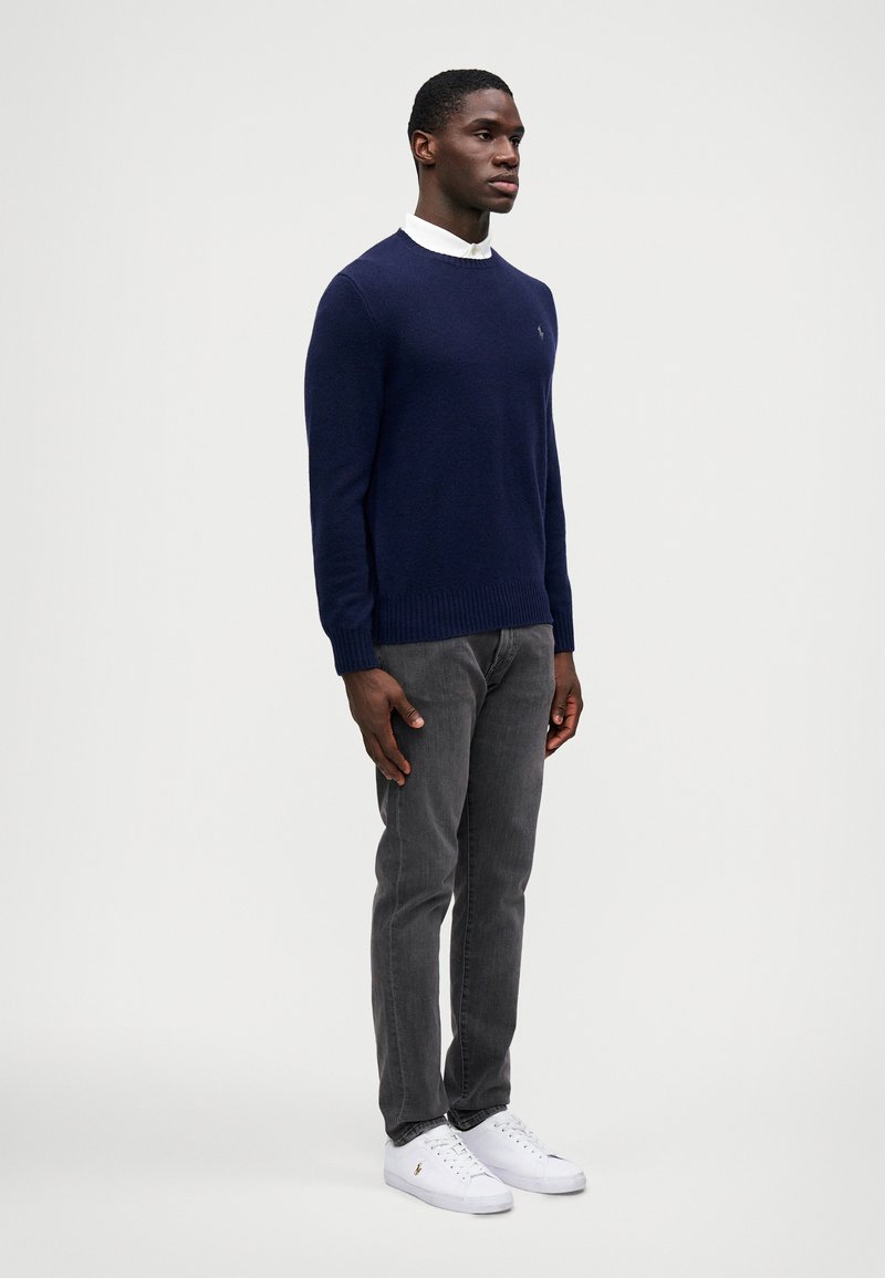 Navy gebreide trui met geribde boorden en zoom, gedragen over een wit overhemd met kraag. Grijze jeans en witte sneakers maken de outfit compleet.