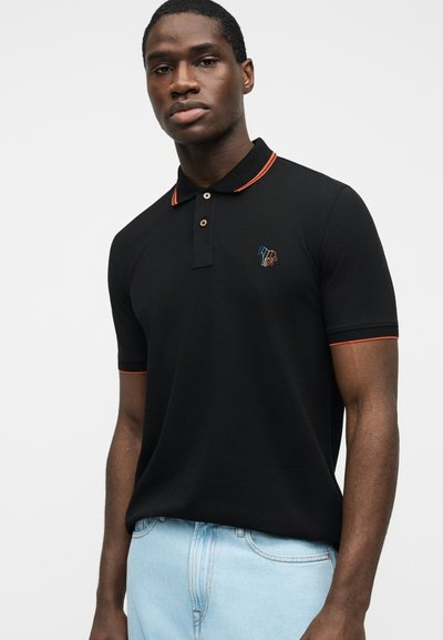 Νέος άνδρας που φοράει μαύρο πόλο με κόκκινη διακόσμηση και ανοιχτόχρωμο μπλε τζιν, όρθιος μπροστά σε απλό λευκό φόντο.