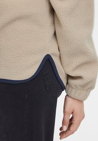 Beige fleece jakke med marineblå kant over sorte bukser, der viser den nederste ærme, sideslids fordybning og en hånd hvilende ved siden.