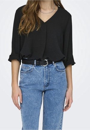 Femme portant un chemisier noir à manches longues avec col en V, rentré dans un jean bleu taille haute avec une ceinture noire et un collier chaîne argenté.