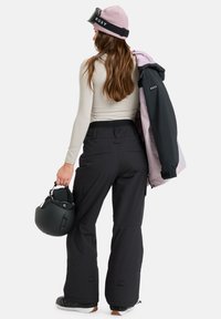 Black snow pants with a relaxed fit, paired with a light pink and black jacket, and a pink beanie. Holding a black ski helmet.