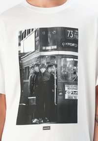 White cotton t-shirt featuring a black and white graphic of five individuals on a bus, with "oasis" label at the bottom.