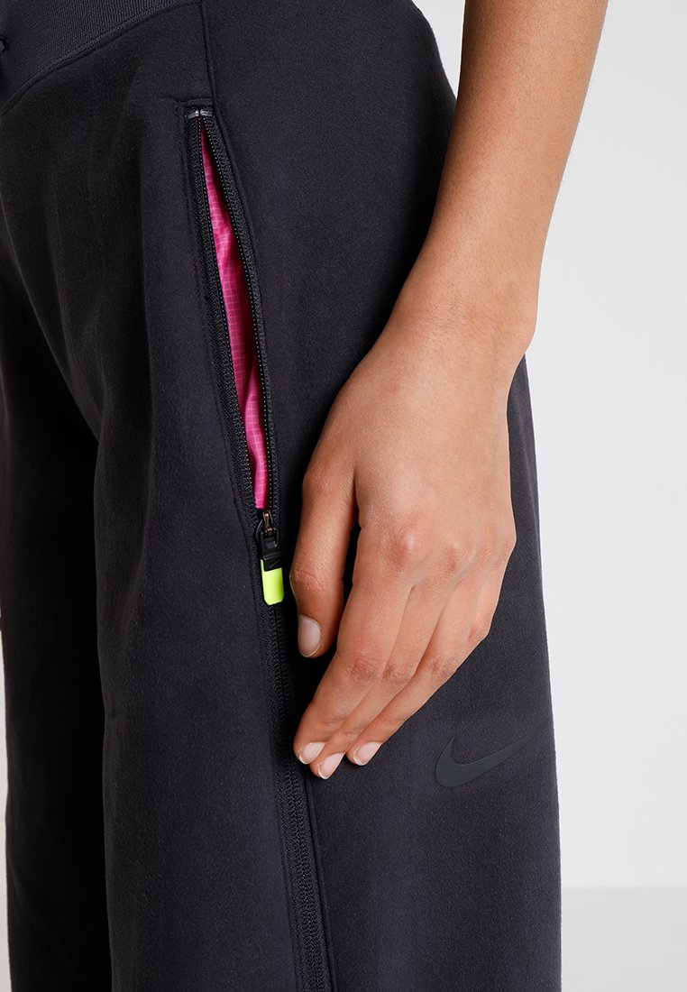 Pantalons de sport noirs avec une doublure rose et une quincaillerie de fermeture éclair visible. Présente un logo Nike en gris foncé sur le côté. Texture lisse.