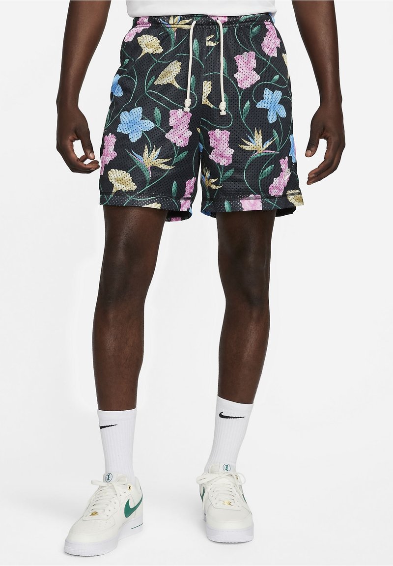 Homme portant un short en mesh noir à imprimé floral rose, bleu et jaune, des chaussettes blanches avec un logo noir, et des baskets blanches avec un swoosh vert.