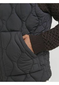 Gepolsterte schwarze Weste mit hohem Kragen, ausgestattet mit einer Seitentasche und gerippten braunen Strickärmeln, die strukturierte Muster auf dem Stoff zeigen.