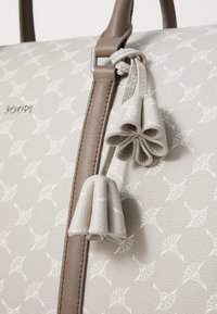 Sac à main gris texturé avec un motif clair répétitif, rehaussé de poignées marron et de pampilles décoratives. Détail du logo en métal visible.