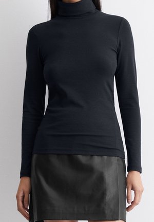 Kobieta w czarnym długim rękawie z golfem i czarnej skórzanej mini spódnicy, stojąca na tle jasnego, jednolitego tła.