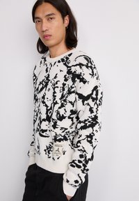 Knitted sweater in white and black, featuring an abstract pattern. Ribbed cuffs and hem, with a tag displaying care instructions at the bottom.