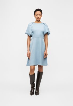 Femme debout devant un arrière-plan uni, portant une robe bleu clair à manches bouffantes et des bottes hautes marron foncé.