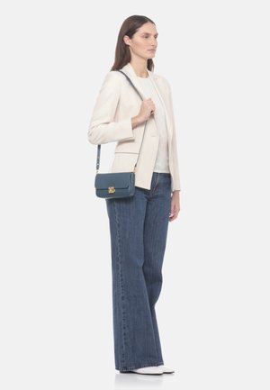 Donna che indossa un blazer crema, top bianco, jeans a zampa larghi blu, scarpe bianche, che tiene una piccola borsa a tracolla blu scuro con chiusura dorata.