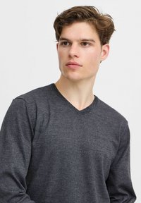 Donkergrijze v-hals trui met een gestructureerd breipatroon, lange mouwen en een aansluitende pasvorm, gedragen door een model met kort haar.