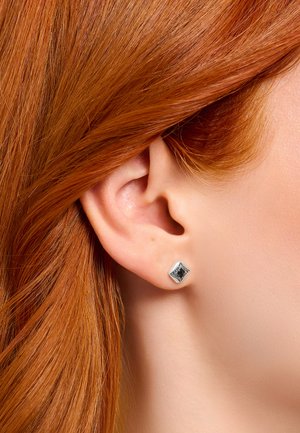 Nahaufnahme eines Ohrs einer rothaarigen Person, die einen kleinen silbernen quadratischen Ohrstecker mit schwarzen und filigranen Details trägt.