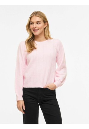 Młoda kobieta o blond włosach, ubrana w jasnoróżowy sweter z długim rękawem i czarne spodnie, uśmiechająca się i patrząca na bok.