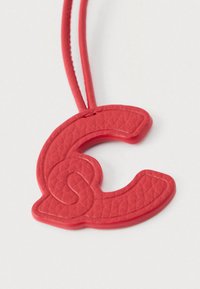 Sarkans teksturēts kulons, kas veidots kā burts C, pievienots atbilstošai sarkanai auklai uz balta fona.