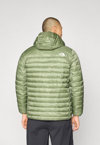 Zelená puffer bunda s kapucňou, prešívaný dizajn, lesklý povrch a logo na hornej časti chrbta. Má voľný strih a elastické manžety.