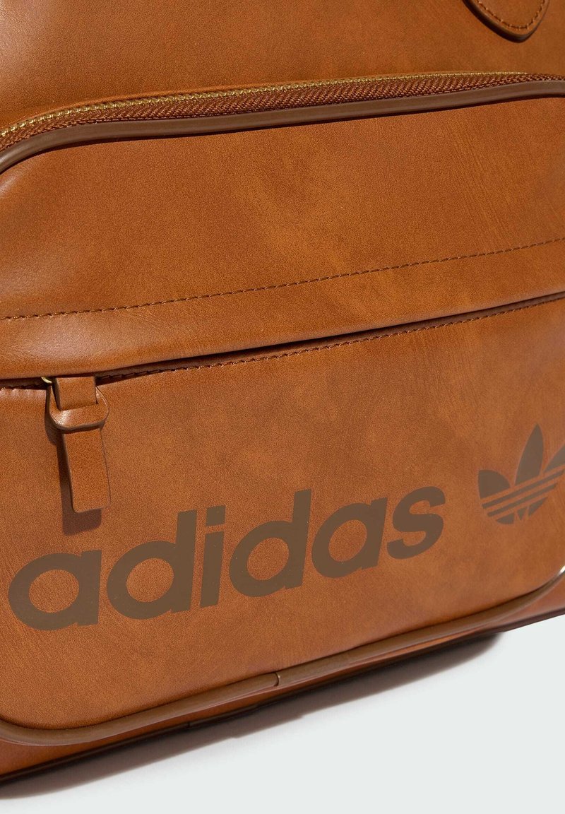 Zaino in pelle marrone con tasca frontale con cerniera, con grande scritta “adidas” e logo trifoglio stampati in marrone scuro.