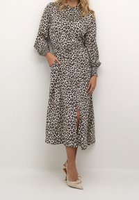 Frau trägt ein langärmeliges, midilanges Kleid mit Leopardenmuster und einem Schlitz vorne, kombiniert mit beigen, slingback Pumps.