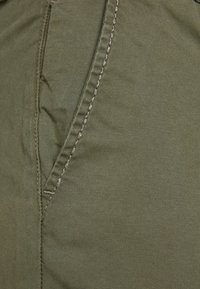 Tessuto verde oliva con una texture liscia, caratterizzato da una tasca laterale cucita e da dettagli di cuciture color crema a contrasto lungo i bordi.